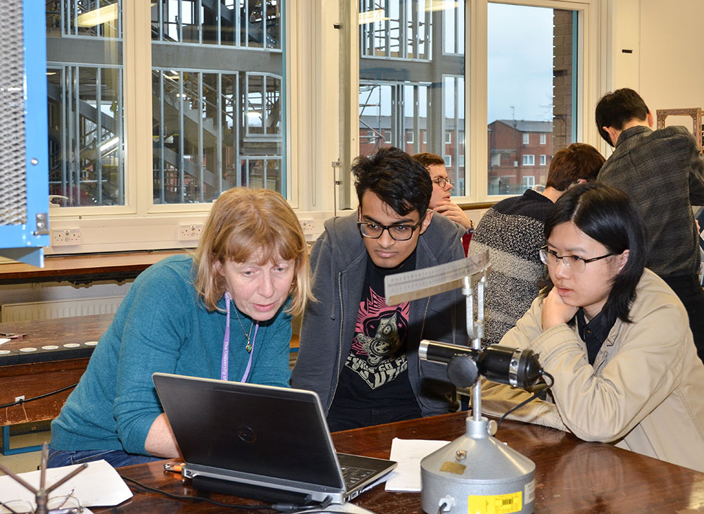 A professor pointing out something on a laptop screen to 2 students in a lab.