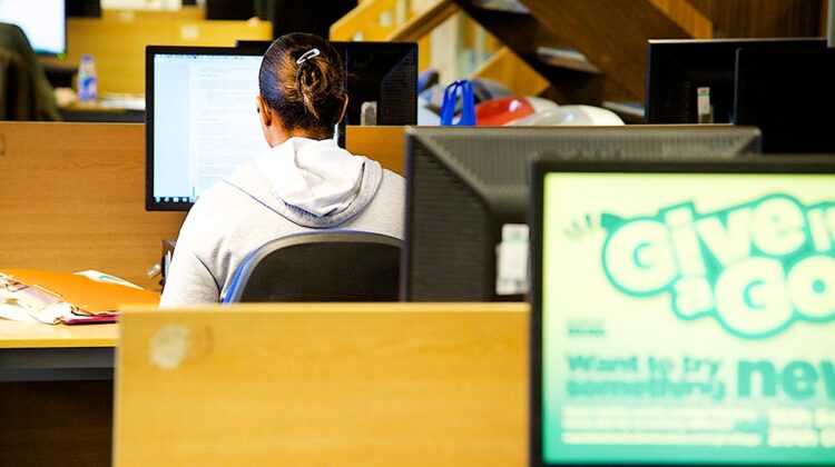 Image of a computer cluster room, with several active screens. You can see one student engaged with their work on screen.