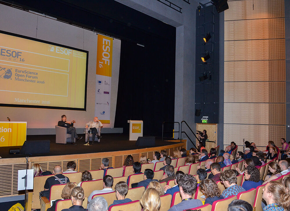 Matthew Cobb and Brian Cox sat on a stage at the Euroscience Open Forum, Manchester 2016, giving a talk to a packed lecture theatre.