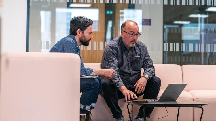 Image of two members of staff sitting on sofas in MECD looking at a laptop screen together.