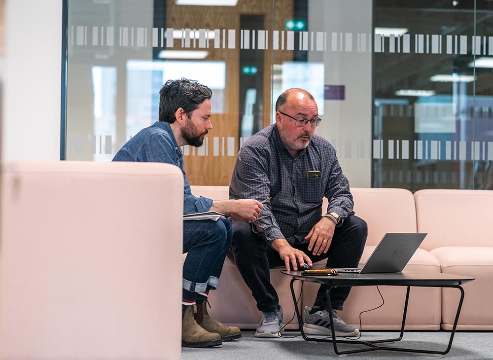 Image of two members of staff sitting on sofas in MECD looking at a laptop screen together.