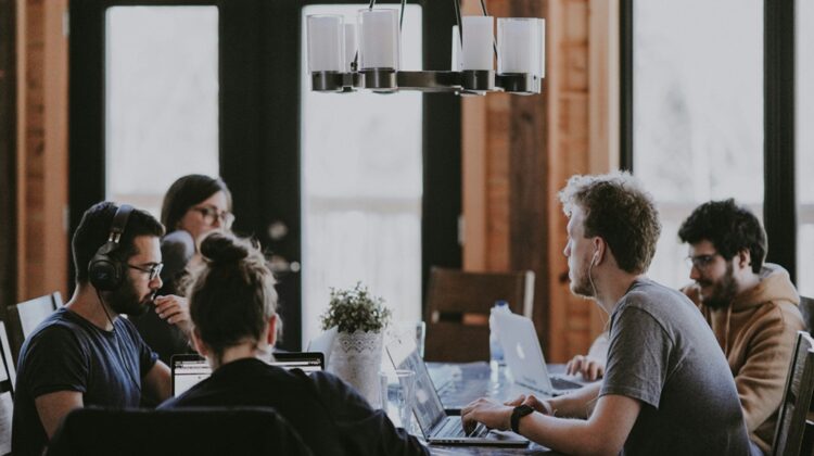 5 people sat around a desk working on laptops
