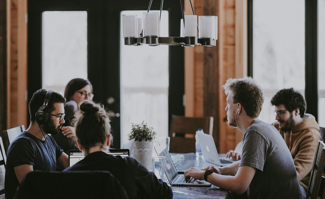 5 people sat around a desk working on laptops