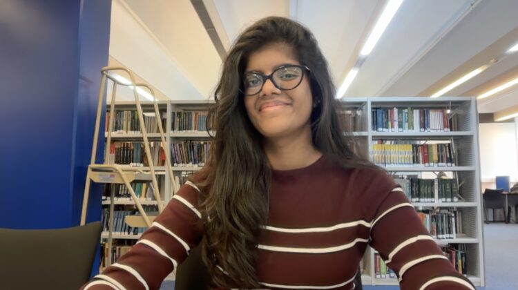A selfie of Rithikha Karunakaran, sat in the library smiling at the camera.